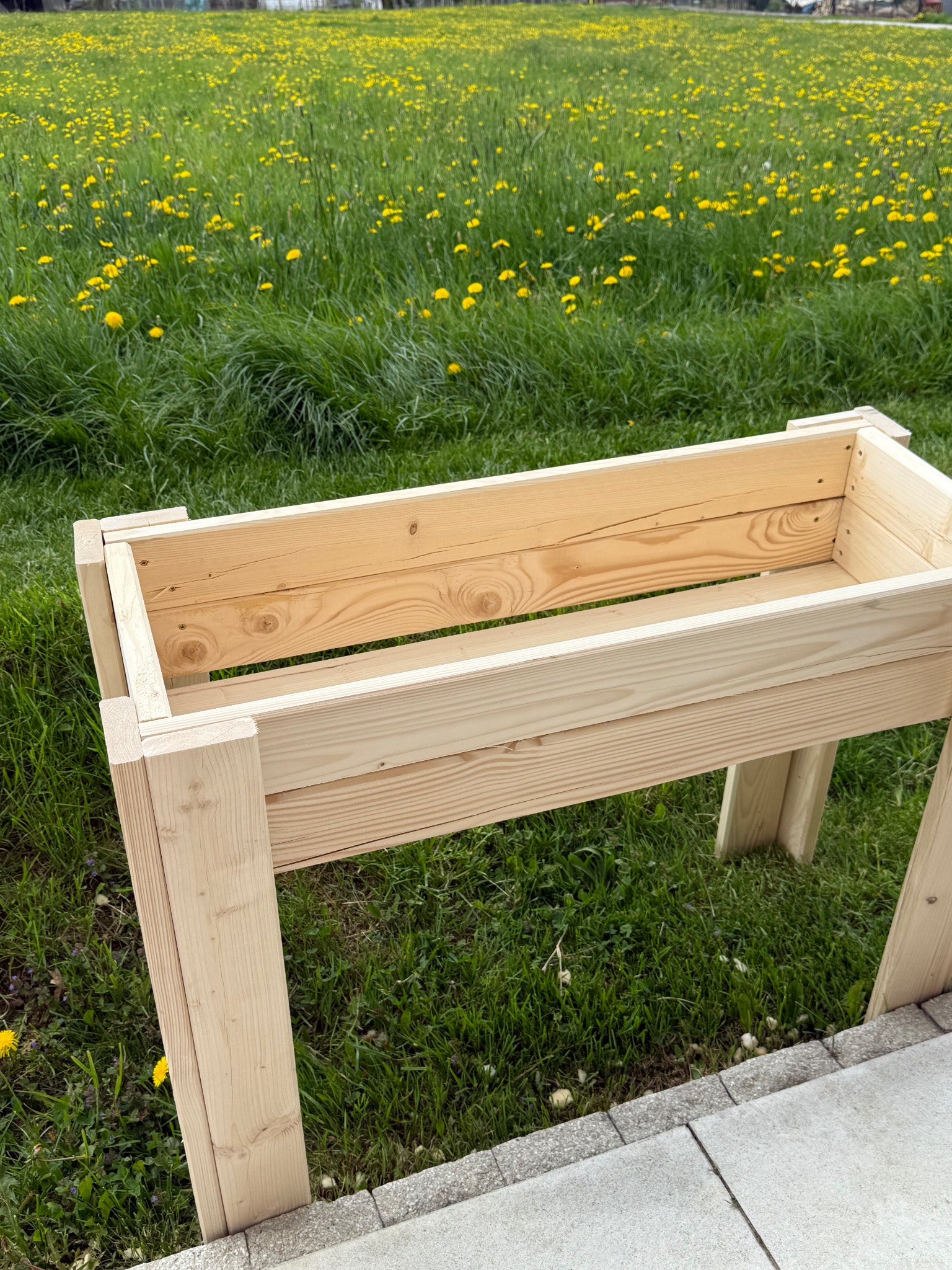 Drewniana skrzynia na nóżkach, wykonana z jasnego drewna, stoi na trawie z żółtymi kwiatami. Skrzynia jest pusta i gotowa do sadzenia roślin.