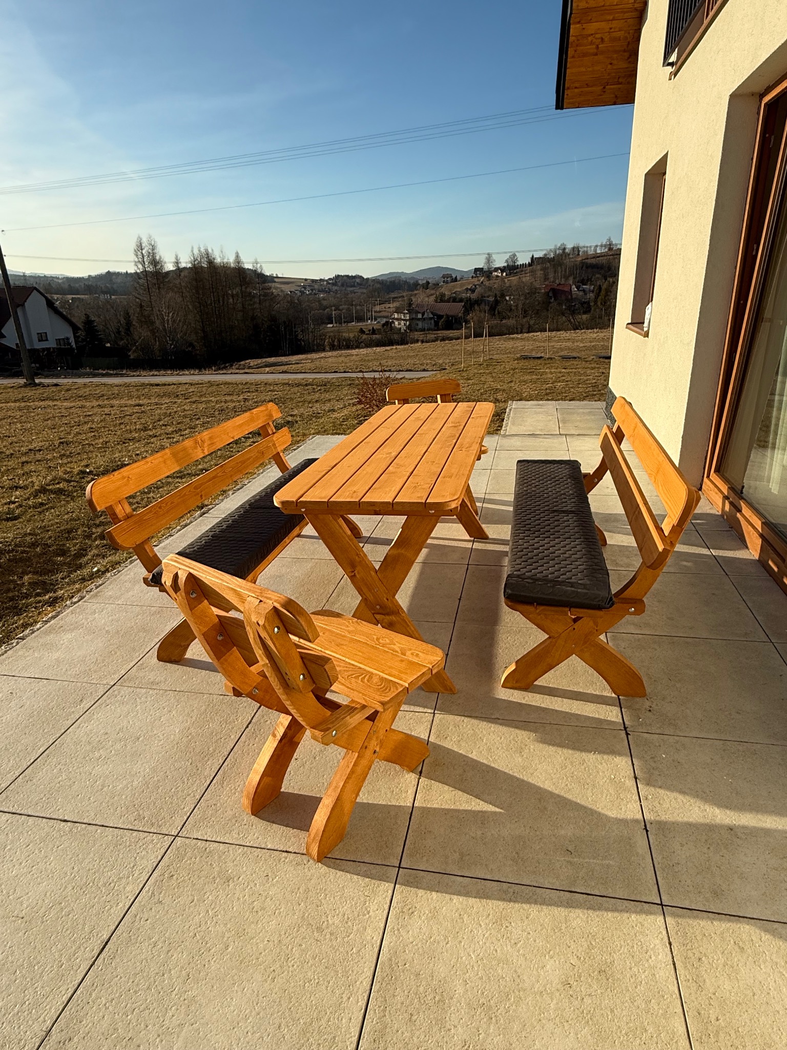 Drewniany zestaw mebli ogrodowych (stół i ławki z poduszkami) na tarasie z widokiem na pagórkowaty krajobraz w słoneczny dzień. Widoczny fragment elewacji domu.