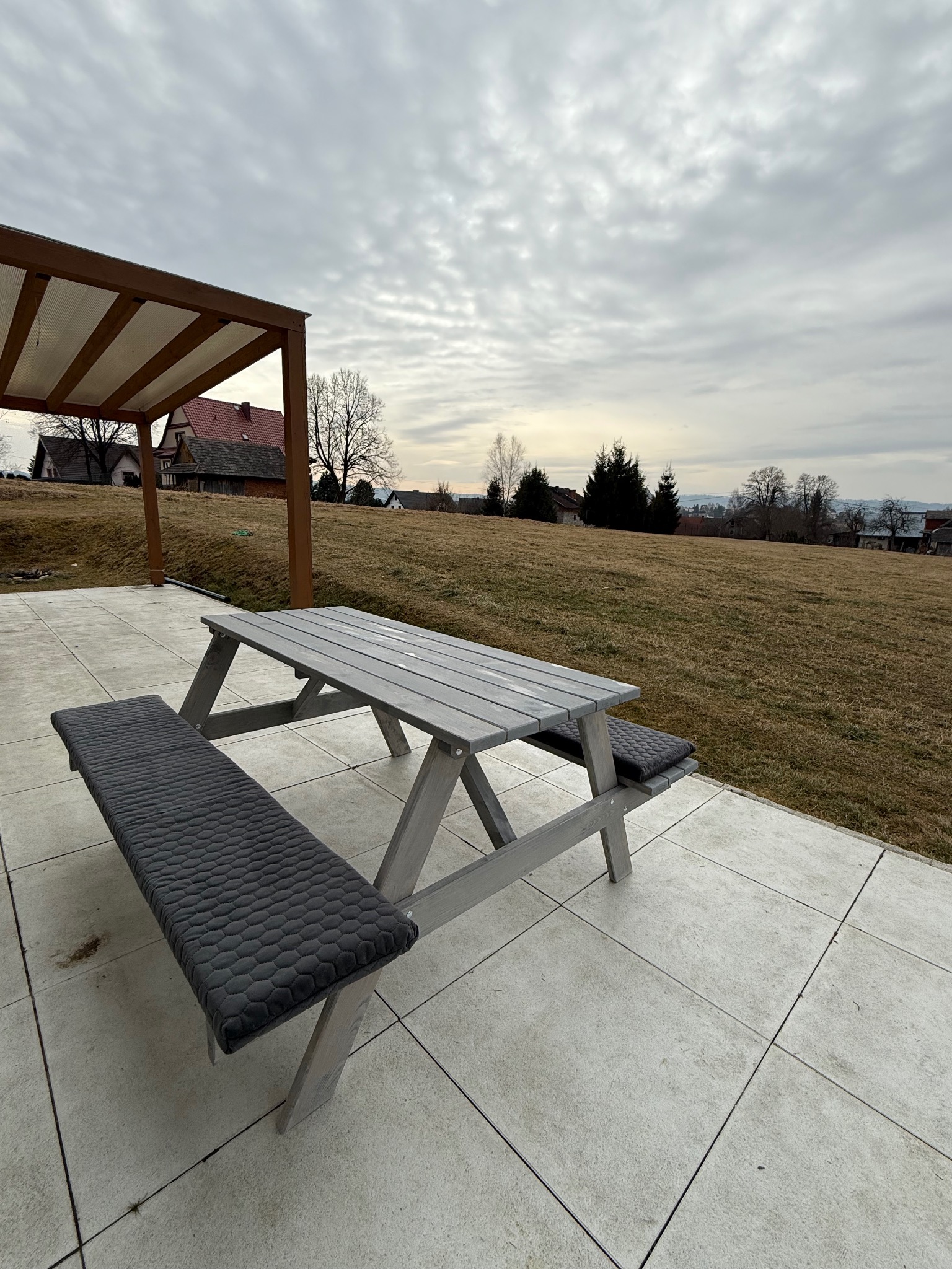 Drewniany stół piknikowy z szarymi poduszkami na tarasie z widokiem na krajobraz. W tle pergola z zadaszeniem i domy. Pochmurne niebo.