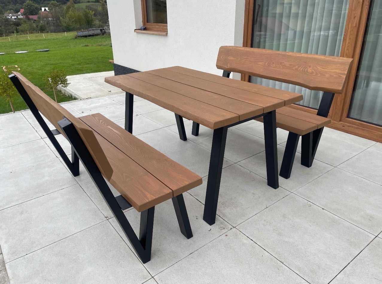 Komplet drewnianych mebli ogrodowych: stół i dwie ławki z oparciami, z czarnymi, metalowymi nogami, na jasnym tarasie, z widokiem na zielony krajobraz.