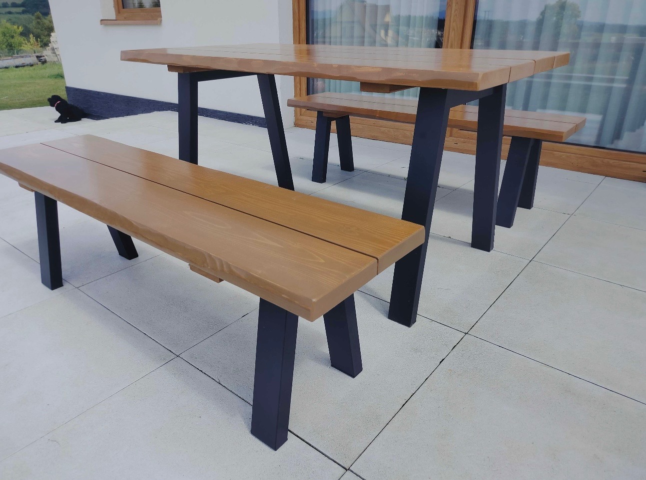 Drewniany stół z ławkami na tarasie z jasnymi płytkami. Blaty w kolorze drewna, nogi czarne. W tle okno i zieleń. Na tarasie czarny pies.