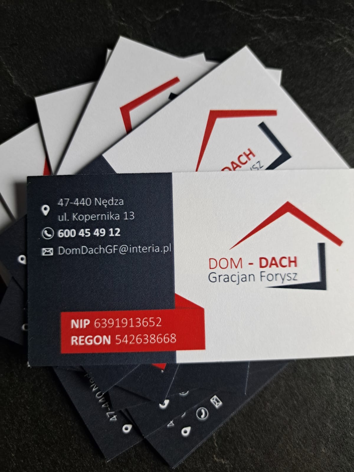 Stos wizytówek firmy dekarskiej Dom-Dach Gracjan Forysz z adresem, numerem telefonu i NIP na ciemnym tle. Minimalistyczny design z czerwono-szarym logo domu.