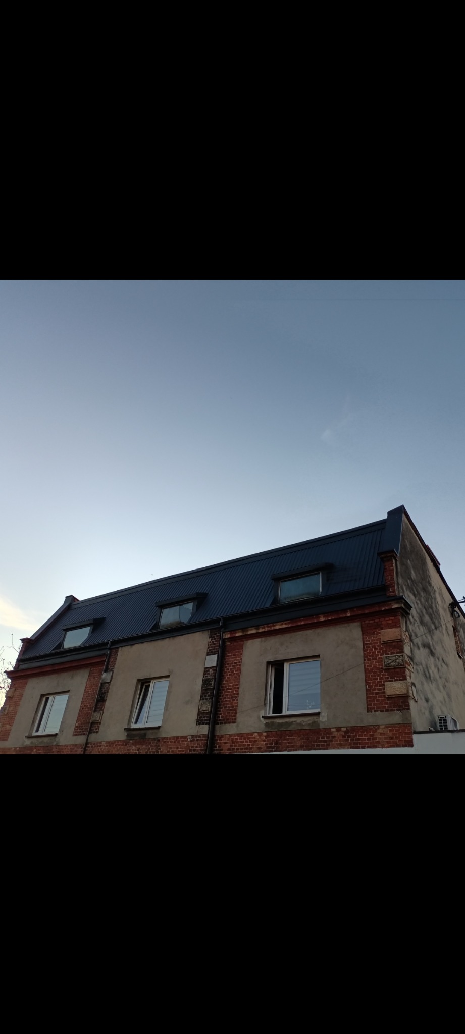 Budynek z ciemnym, metalowym dachem i oknami dachowymi, widziany pod kątem. Ceglane elementy dekoracyjne na elewacji. Błękitne niebo w tle.
