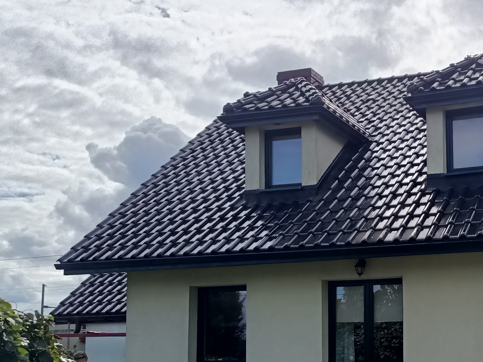 Czarny dach z dachówki ceramicznej na jasnym domu z oknami w ciemnych ramach, widok z dołu na tle pochmurnego nieba. Widoczne rynny i komin.