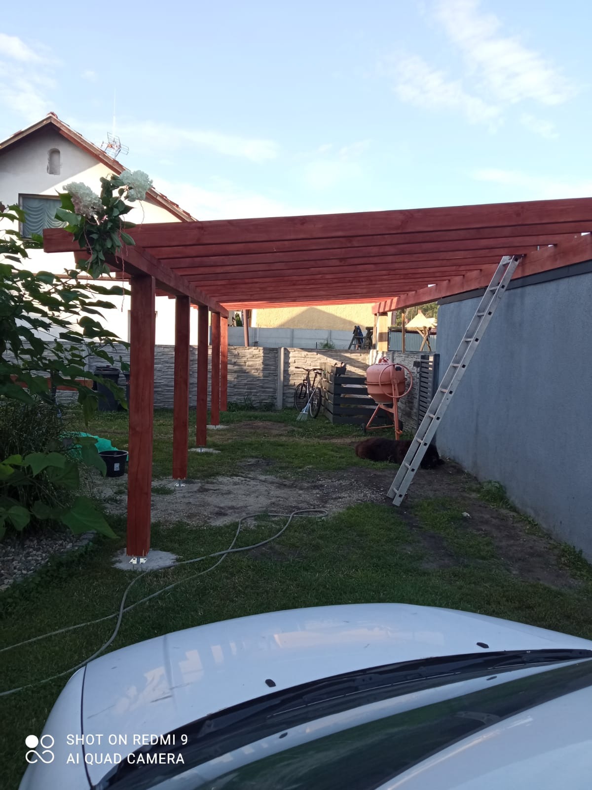 Drewniana pergola w ogrodzie, świeżo pomalowana na brązowo, z widoczną drabiną i betoniarką w tle. Częściowo widoczny samochód na pierwszym planie.