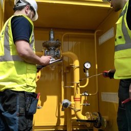 PM Instal - Marcin Piórkowski - Dwóch gazowników w żółtych kamizelkach sprawdza instalację gazową w żółtej obudowie, jeden z miernikiem, drugi z notatnikiem, kontrola zaworów i rur.