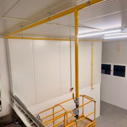 PM Instal - Marcin Piórkowski - Żółta instalacja gazowa pod sufitem w hali, widoczna z podnośnika. Rury zamocowane do sufitu, ściany z paneli. Widok z góry na podnośnik Haulotte Optimum 8.