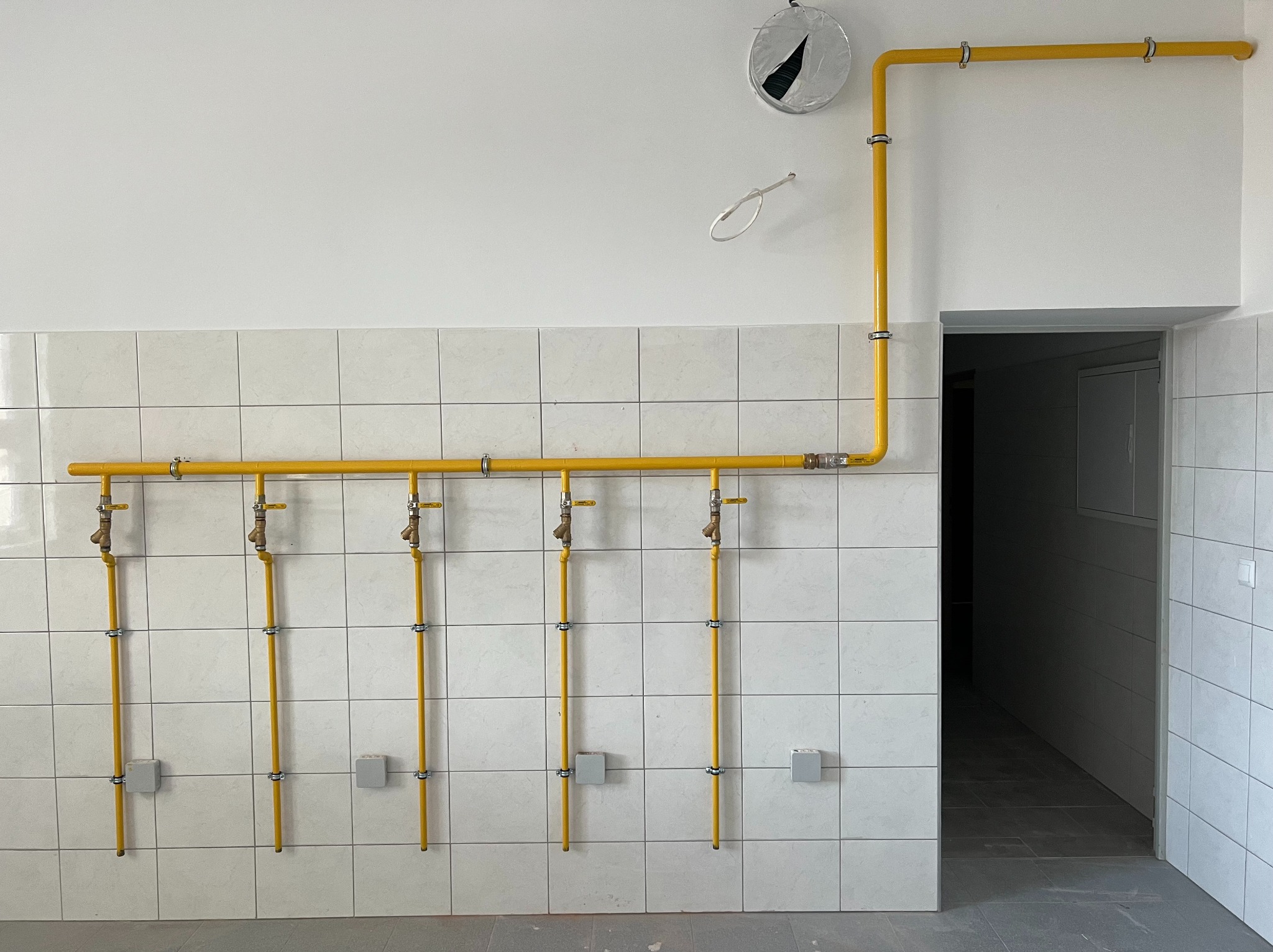 Żółte rury gazowe z kranami wzdłuż kafelkowanej ściany w kuchni placówki szkolnej, z widocznymi podłączeniami do kuchenek palnikowych i taboretowych.