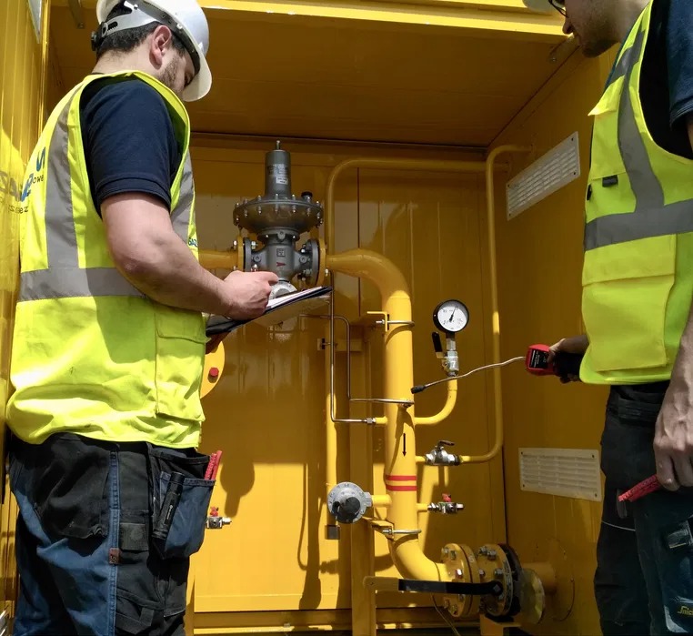 Dwóch gazowników w żółtych kamizelkach sprawdza instalację gazową w żółtej obudowie, jeden z miernikiem, drugi z notatnikiem, kontrola zaworów i rur.
