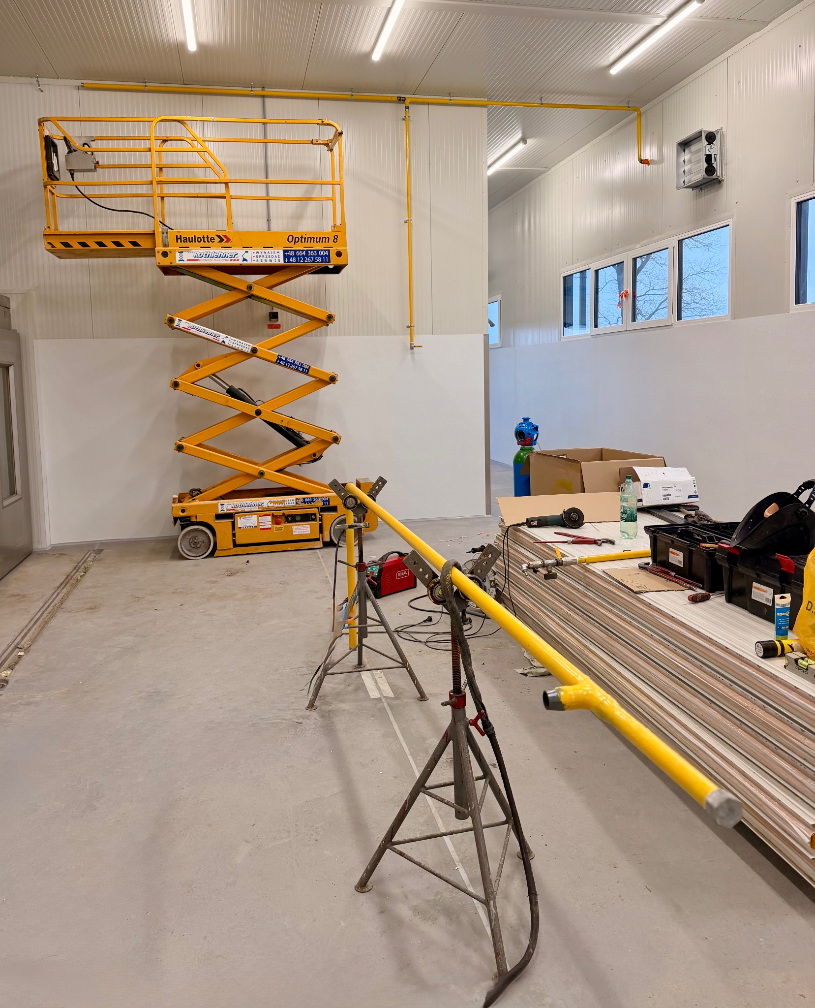 Żółta instalacja gazowa w hali, widoczna z podnośnika nożycowego. Rury na stojakach, narzędzia i materiały na podłodze. Oświetlenie jarzeniowe, białe ściany z paneli.