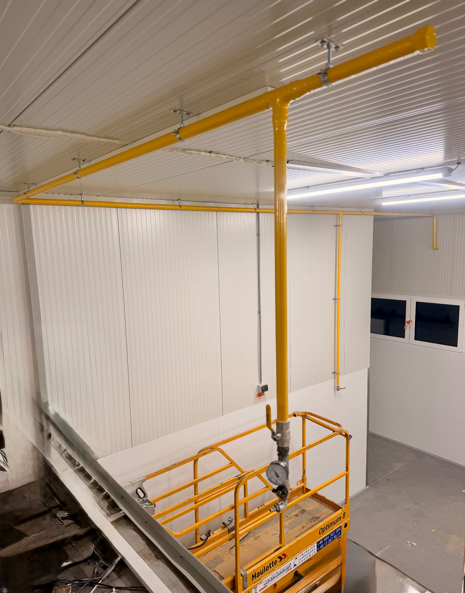 Żółta instalacja gazowa pod sufitem w hali, widoczna z podnośnika. Rury zamocowane do sufitu, ściany z paneli. Widok z góry na podnośnik Haulotte Optimum 8.