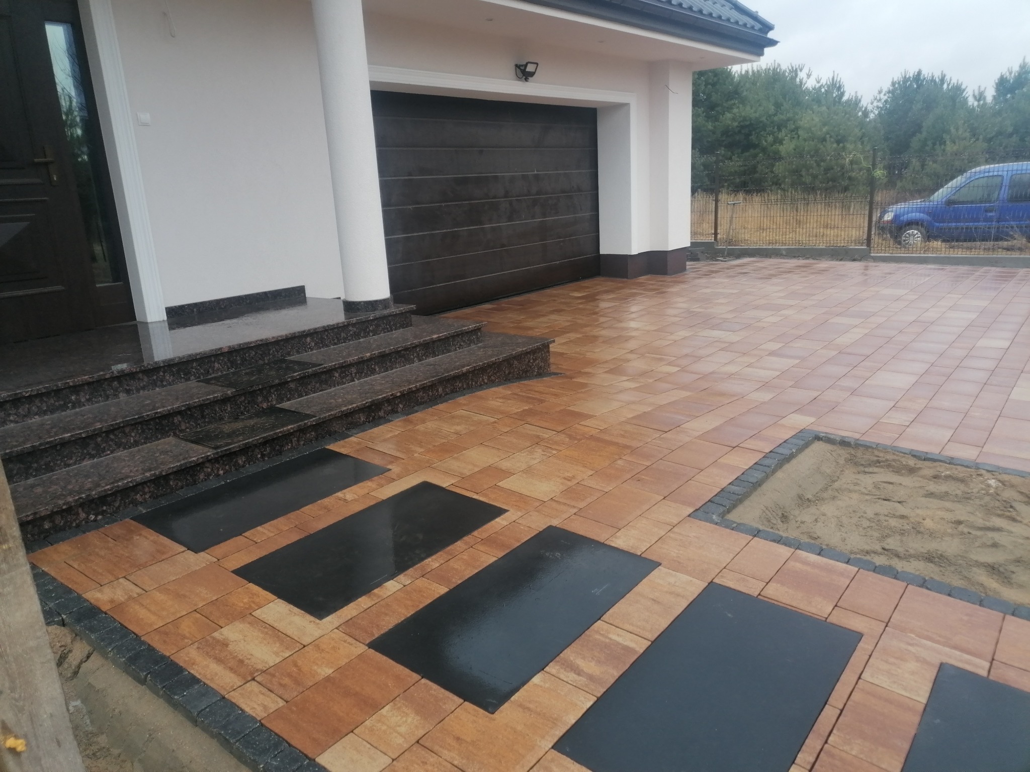 Nowoczesny podjazd z kostki brukowej w odcieniach brązu i czerni, z elementami dekoracyjnymi w postaci prostokątnych płyt. Schody wejściowe z ciemnego granitu.