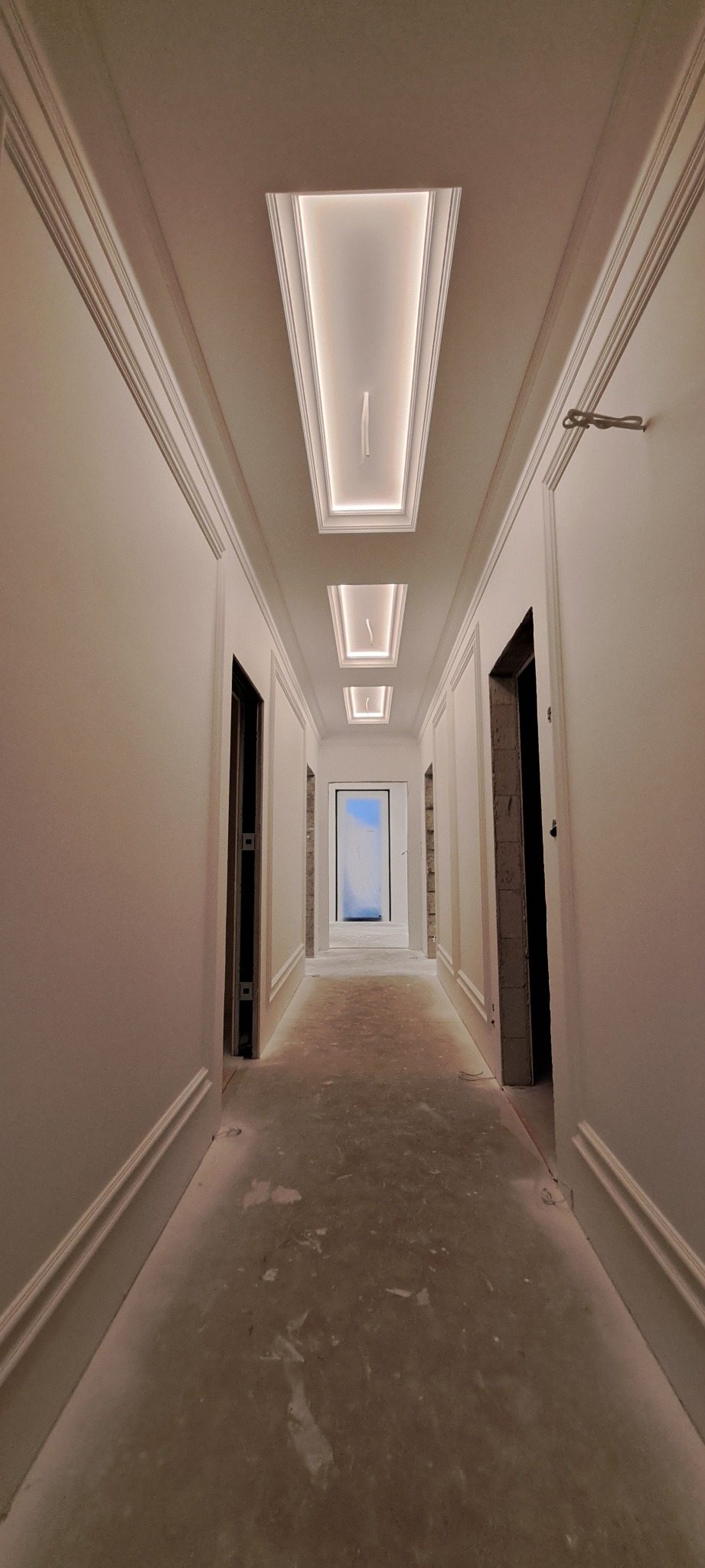 Korytarz po remoncie: białe ściany z dekoracyjnymi listwami, sufit z podświetleniem LED w prostokątnych wnękach. Widoczne niedokończone ościeżnice i zabezpieczone drzwi.