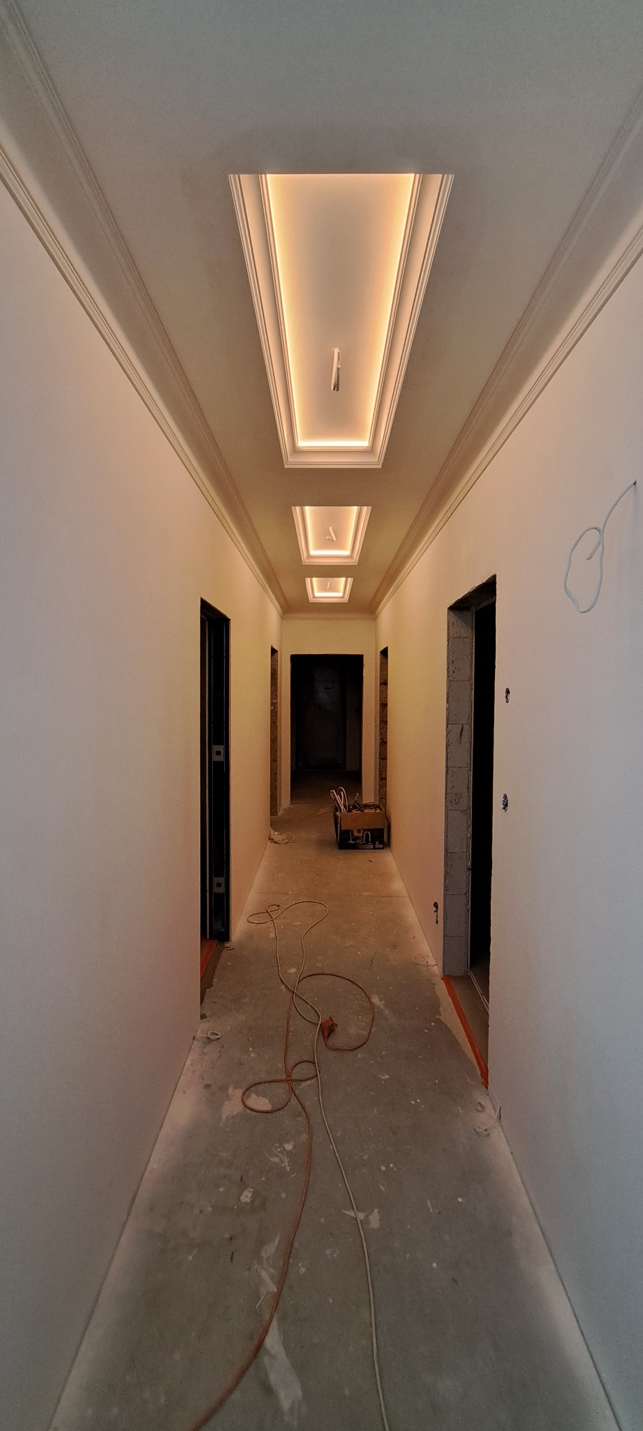 Korytarz w trakcie wykończenia: świeżo pomalowane ściany, dekoracyjne listwy przysufitowe i podświetlany sufit kasetonowy. Widoczne przewody na podłodze.