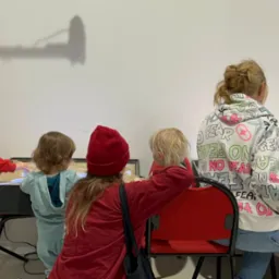 Warsztaty artystyczne dla dzieci: grupa maluchów tworzy prace plastyczne przy podświetlanym stole pod okiem opiekunów. Widoczne materiały plastyczne i oświetlenie studyjne.