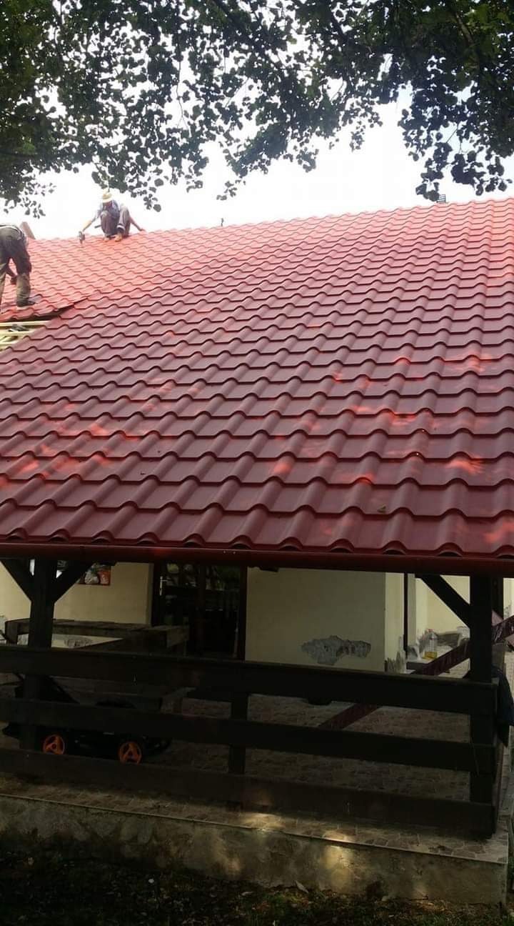 Czerwony dach z blachodachówki, widok z dołu na budynek, dwóch dekarzy pracuje na górze. Częściowo widoczna drewniana konstrukcja.