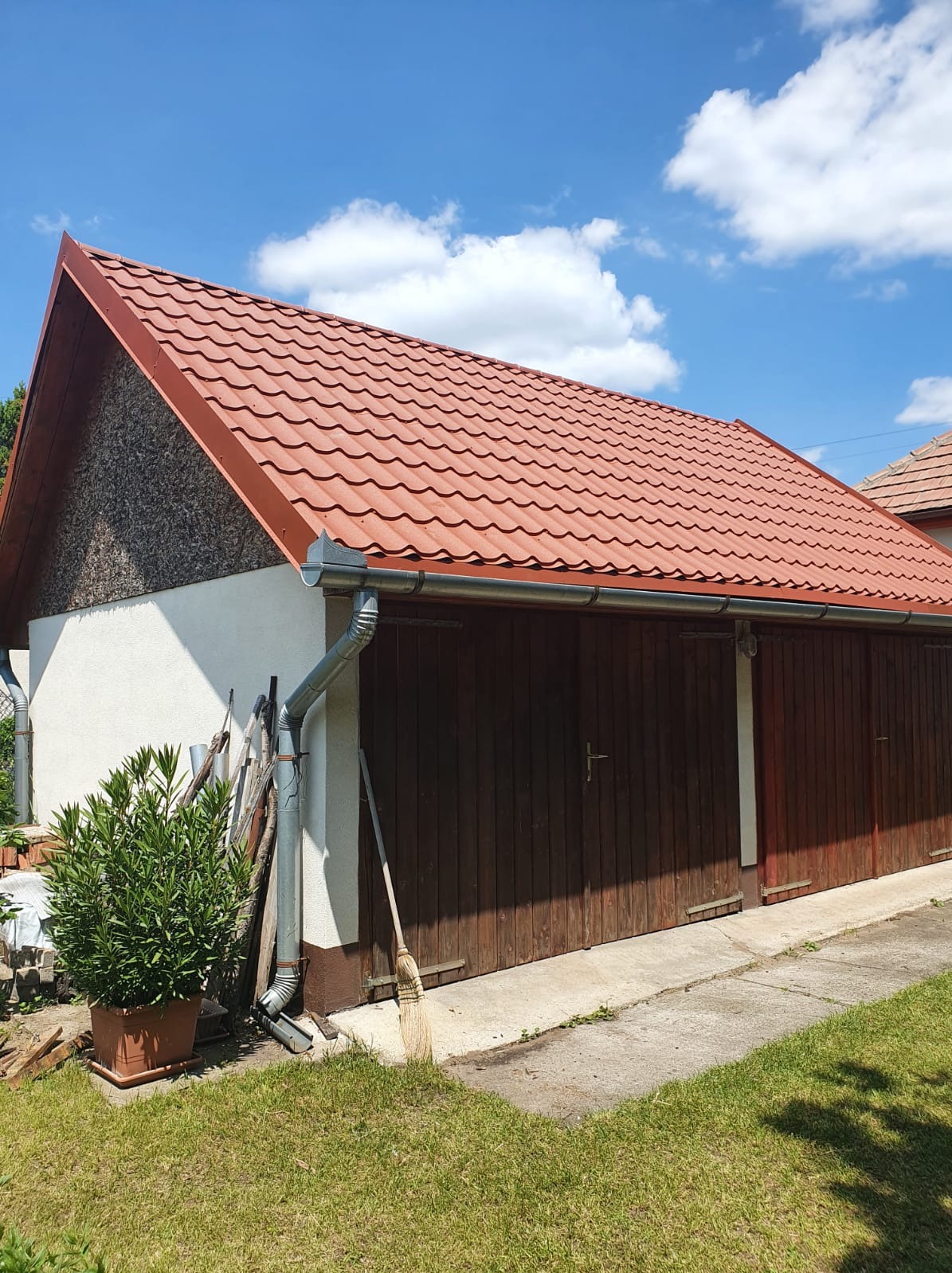 Czerwony dach z blachodachówki na budynku gospodarczym z drewnianymi wrotami garażowymi i rynną spustową w słoneczny dzień.