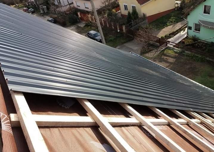 Szary dach z blachy trapezowej w trakcie montażu, widoczna więźba dachowa i membrana. W tle domy jednorodzinne i zieleń.