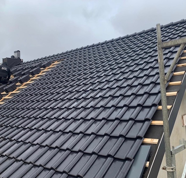 Układanie czarnej dachówki na dachu domu. Widoczny fragment rusztowania i dekarz w trakcie pracy. Dachówka błyszcząca, ułożona w rzędach, na tle pochmurnego nieba.