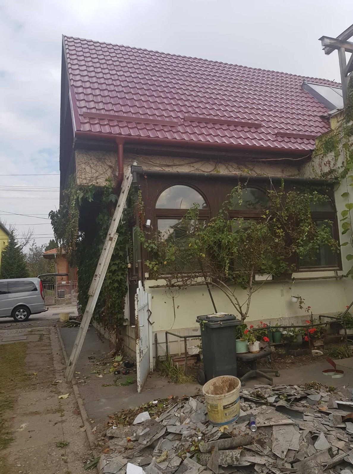 Dom z nowym, bordowym dachem z blachodachówki trapezowej. Przy ścianie oparta drabina, w tle widoczny samochód. Na chodniku sterta starych dachówek.