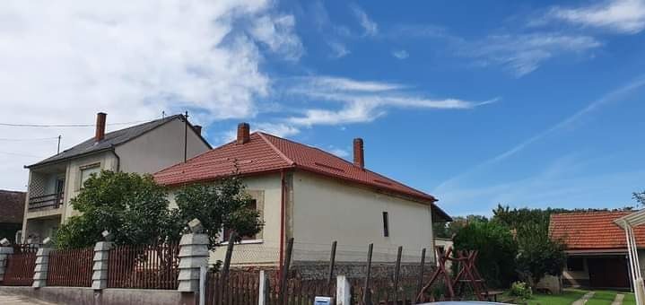 Dom jednorodzinny z czerwonym dachem i ceglanym kominem na tle błękitnego nieba z chmurami. Widoczny fragment ogrodzenia i zieleń drzew.