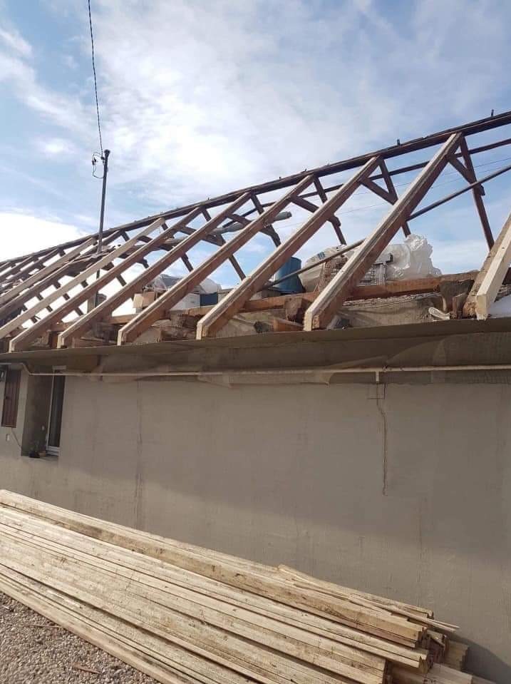 Wymiana więźby dachowej w trakcie: widoczne drewniane elementy konstrukcyjne na tle błękitnego nieba, z zapasem desek u dołu zdjęcia.