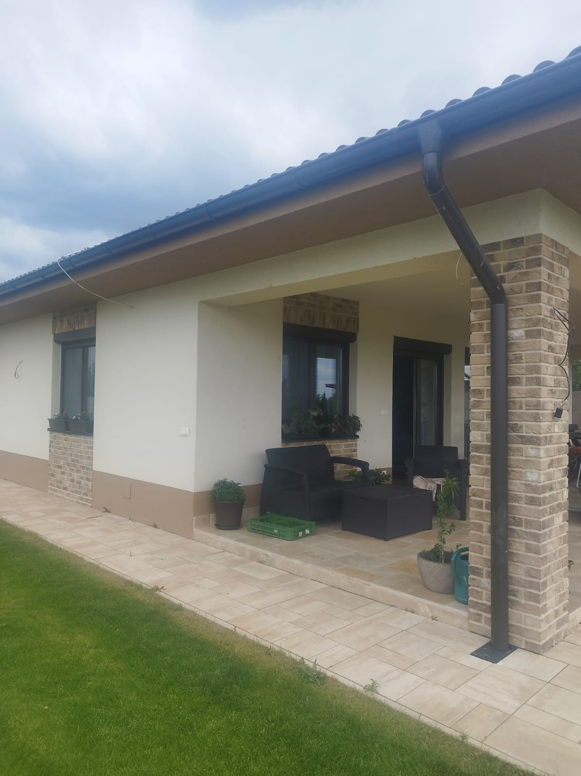 Elewacja domu z widocznymi rynnami w kolorze grafitowym, wykończona cegłą klinkierową i jasnym tynkiem. Na pierwszym planie trawnik i taras z meblami ogrodowymi.