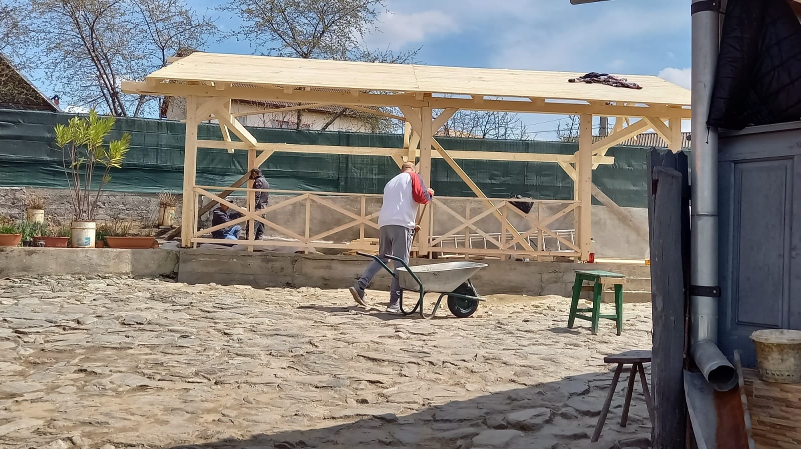 Budowa drewnianej altany z jasnego drewna. W tle zielona siatka i budynek. Na pierwszym planie mężczyzna z taczką na brukowanym podwórku.