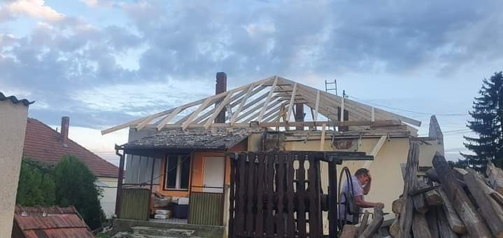 W trakcie budowy więźby dachowej domu jednorodzinnego. Na pierwszym planie drewno budowlane, w tle widoczny dekarz przy pracy oraz drabina oparta o konstrukcję dachu.