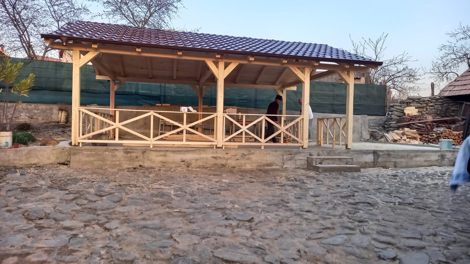 Drewniana altana z brązowym dachem z blachodachówki, w trakcie budowy. Widoczne elementy konstrukcyjne i fragmenty ogrodzenia. Dwóch mężczyzn przy pracy.