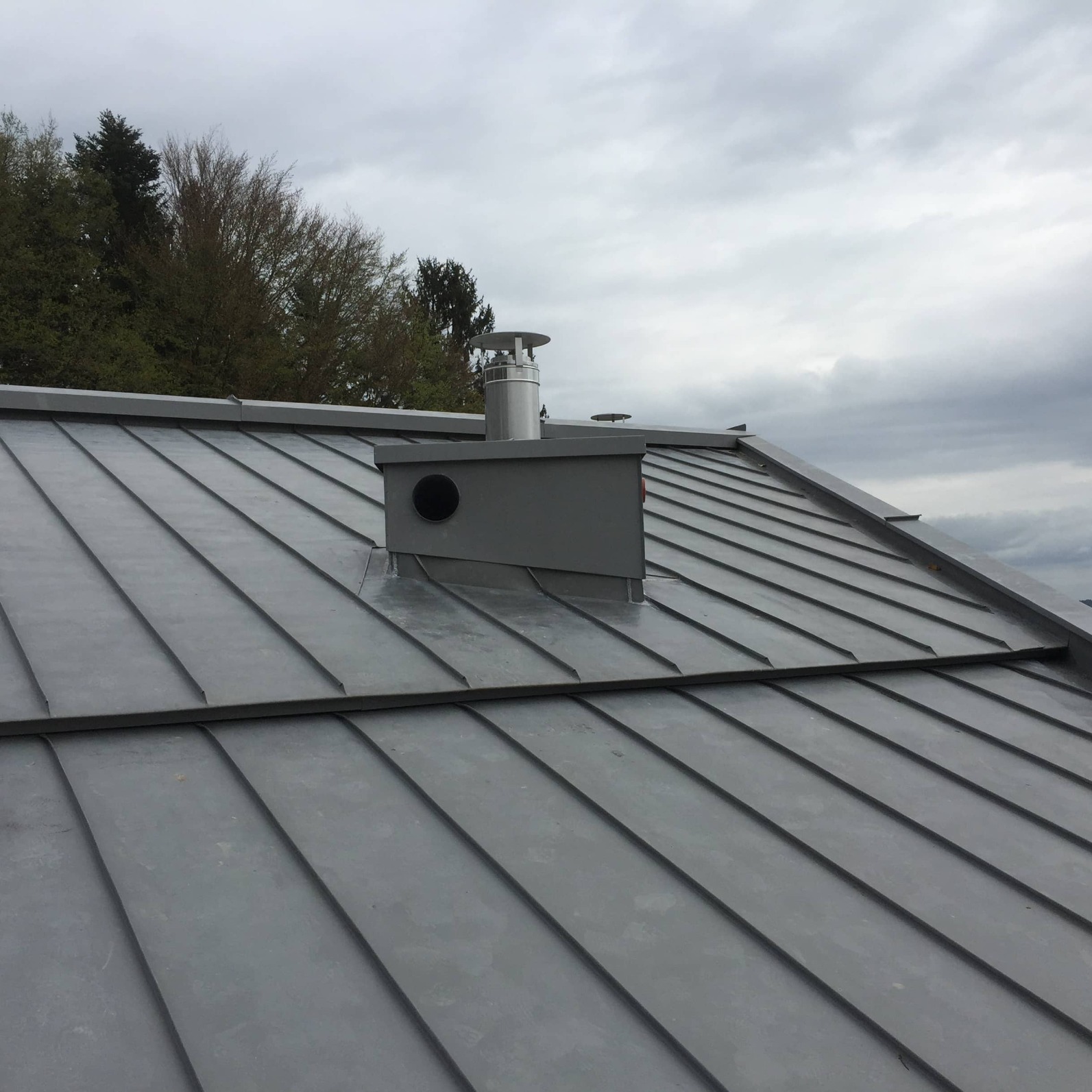 Szary dach z blachy na rąbek stojący z kominem i odpowietrznikiem na tle pochmurnego nieba i drzew. Widok z góry.
