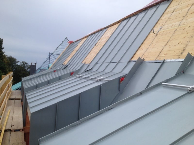 Szary dach z blachy na budynku w trakcie budowy, widoczne deski i zabezpieczenia. Ujęcie z dołu ukazuje detale konstrukcji i materiałów.