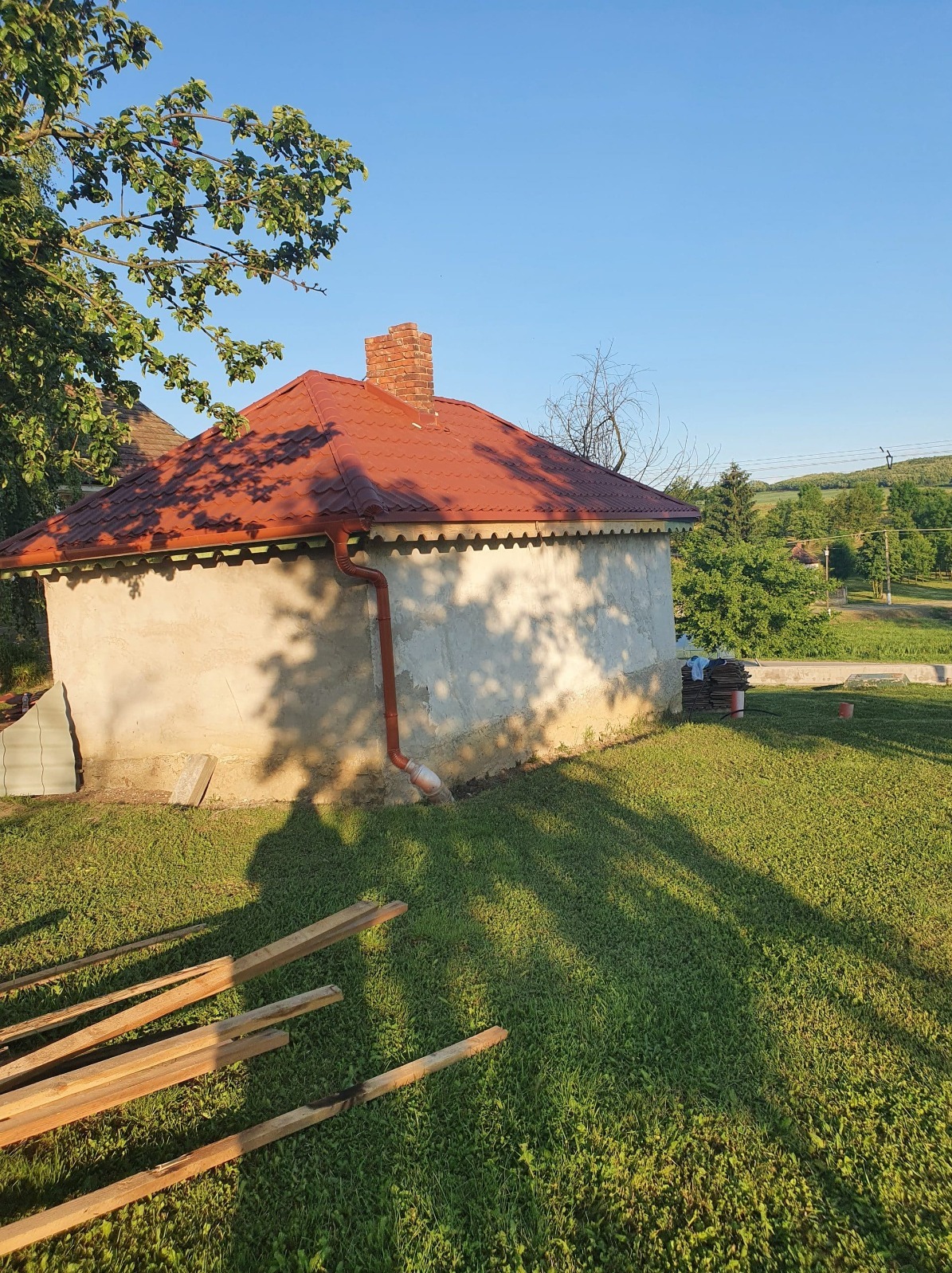 Niski budynek z czerwoną dachówką, ceglanym kominem i orynnowaniem. W tle zielone wzgórza i błękitne niebo. Na pierwszym planie drewniane elementy konstrukcyjne.