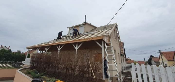 Dwóch dekarzy pracuje na dachu domu jednorodzinnego, widoczna drabina oparta o ścianę, częściowo zdemontowane pokrycie dachowe i przygotowana więźba.