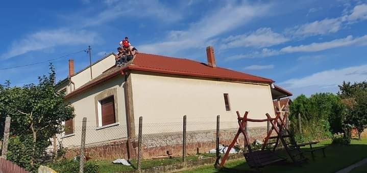 Dom z nowym, czerwonym dachem i rynnami. Na dachu grupa osób, prawdopodobnie ekipa dekarska podczas pracy. Błękitne niebo w tle.