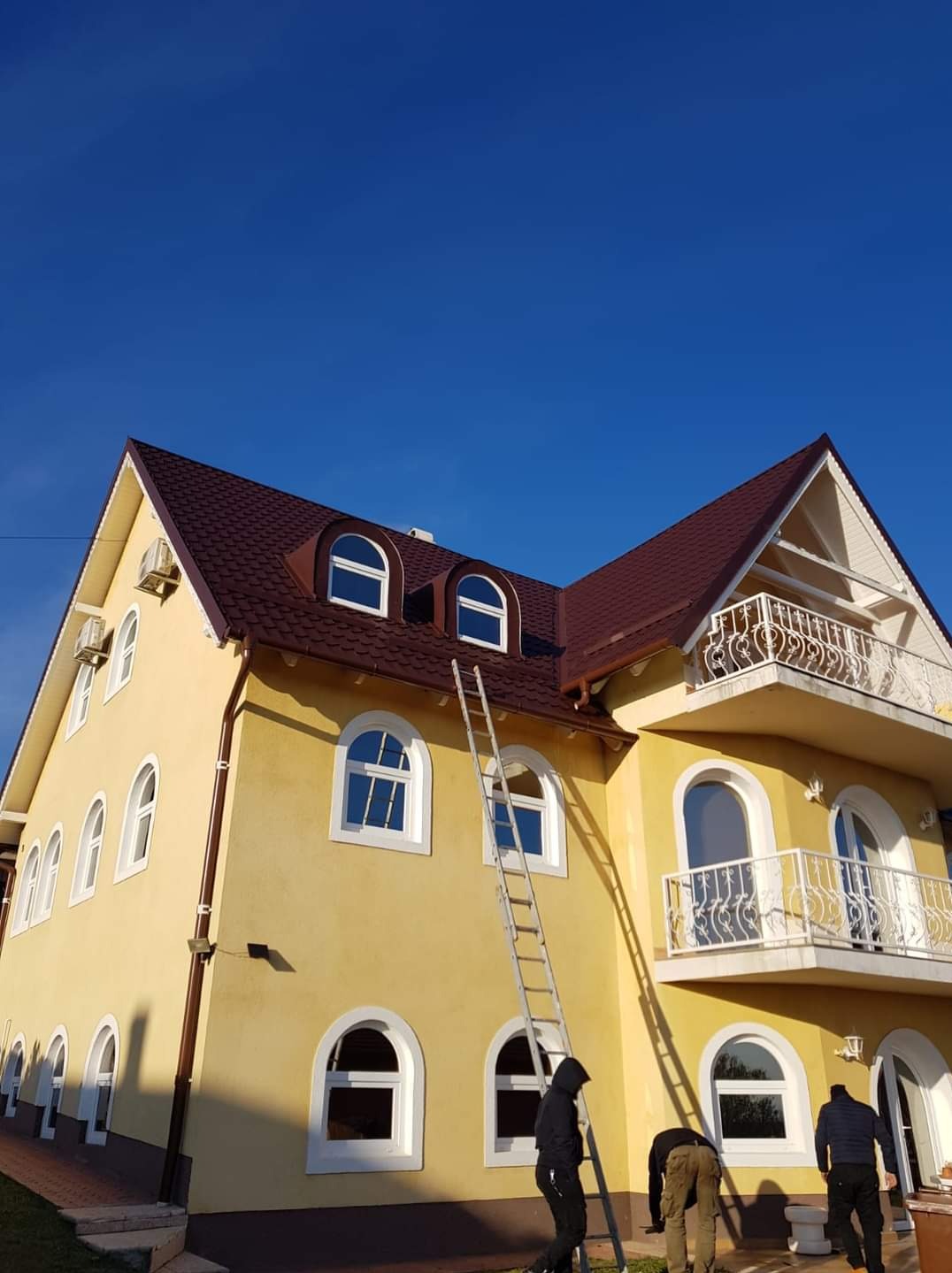 Żółty dom z brązowym dachem krytym dachówką, drabina oparta o dach, pracownicy na dole. Ujęcie z dołu, błękitne niebo w tle.