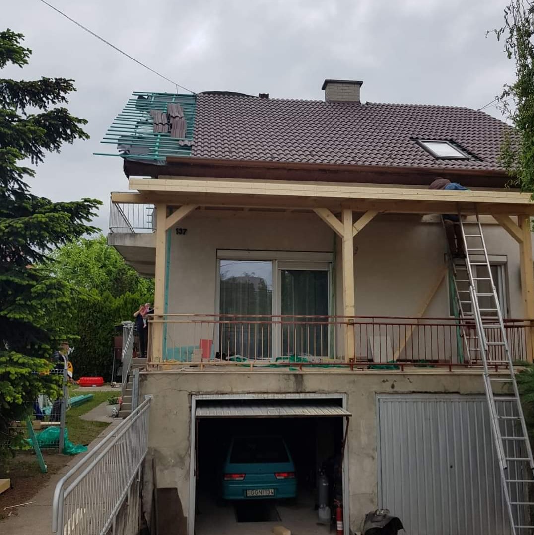 Dom jednorodzinny w trakcie wymiany pokrycia dachowego. Widoczna więźba i częściowo zdjęte stare dachówki. Na dachu pracownik. W tle pochmurne niebo.