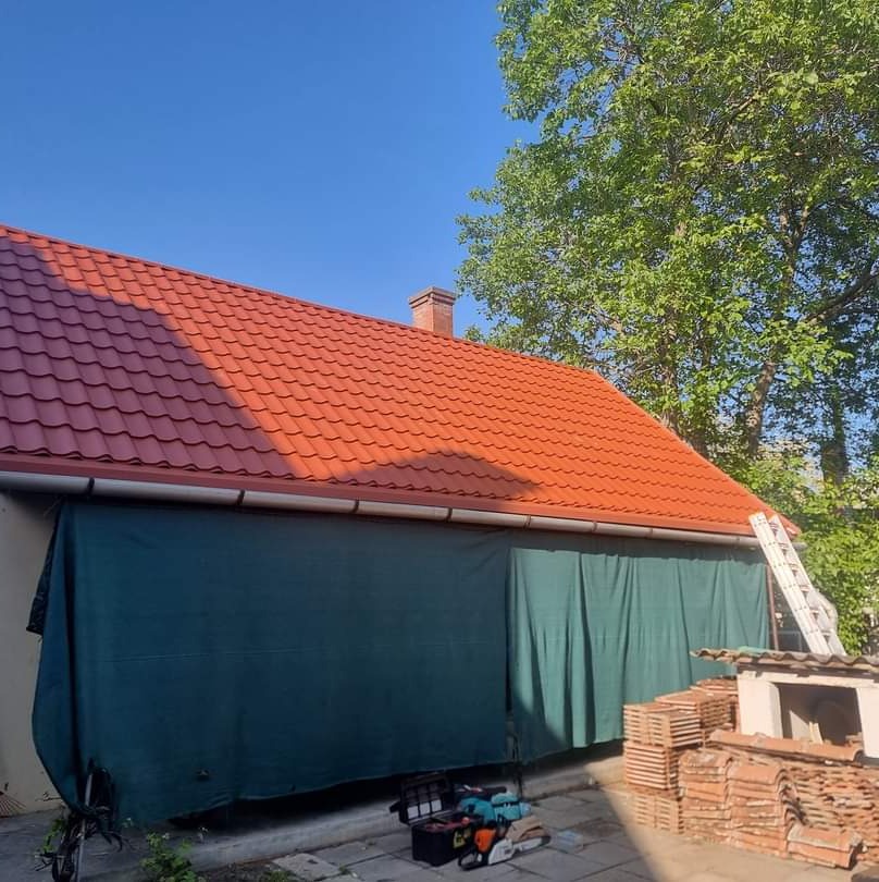 Czerwony dach z blachodachówki, widoczny komin i rynny. Pod dachem rozwinięta plandeka, przy ścianie drabina i narzędzia. Jasne niebo, drzewa w tle.