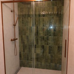 Twoje Nowe Miejsce - Kabina prysznicowa z przesuwnymi drzwiami, wykończona zielonymi, pionowo ułożonymi płytkami. Armatura w kolorze miedzi. Minimalistyczny design.