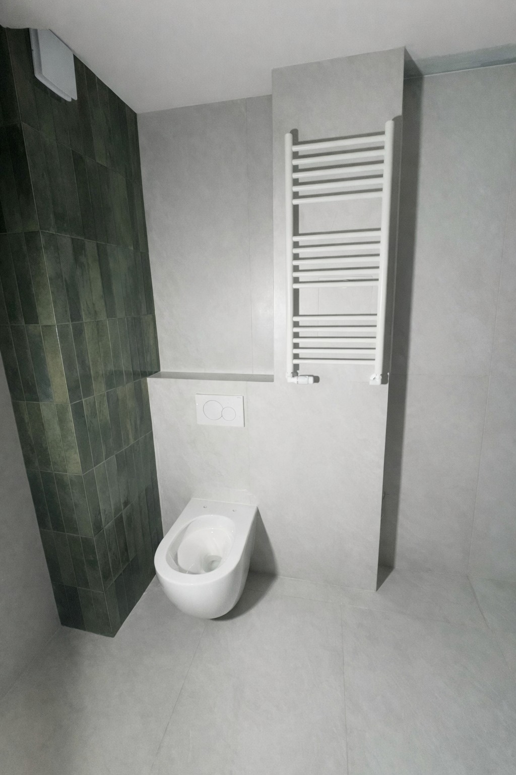 Nowoczesna łazienka z wiszącą toaletą, grzejnikiem drabinkowym i płytkami w odcieniach szarości oraz zieleni. Minimalistyczny design z akcentem kolorystycznym.