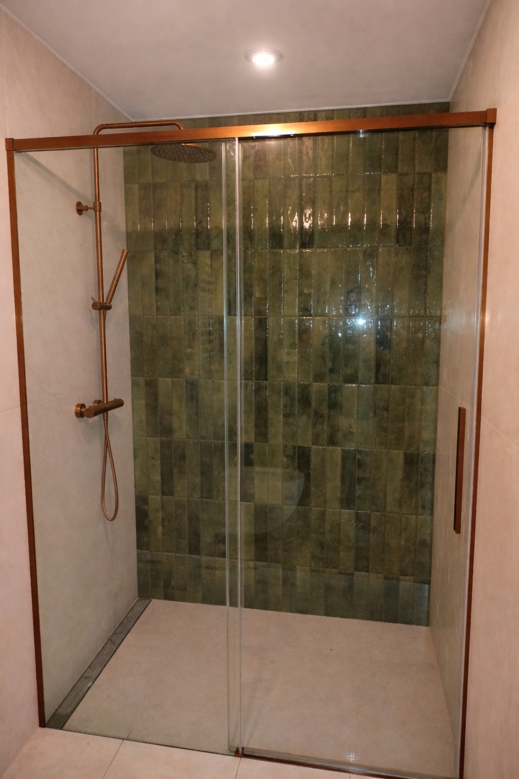 Kabina prysznicowa z przesuwnymi drzwiami, wykończona zielonymi, pionowo ułożonymi płytkami. Armatura w kolorze miedzi. Minimalistyczny design.