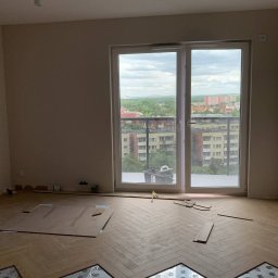 Marcin Petrykowski Wemar-Rem - Układanie parkietu w jodełkę w jasnym pokoju z widokiem na miasto. Częściowo ułożona podłoga, widoczna folia izolacyjna. Praca w toku, naturalne światło.