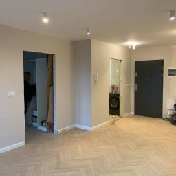 Marcin Petrykowski Wemar-Rem - Jasne panele ułożone w jodełkę w przestronnym wnętrzu z beżowymi ścianami i widocznymi otworami drzwiowymi. W tle pralka i osoba w pomieszczeniu.