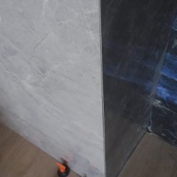 Marcin Petrykowski Wemar-Rem - Ułożone płytki podłogowe imitujące drewno, z użyciem systemu poziomowania płytek, przy narożniku ściany wyłożonej szarą i ciemną płytką imitującą marmur.