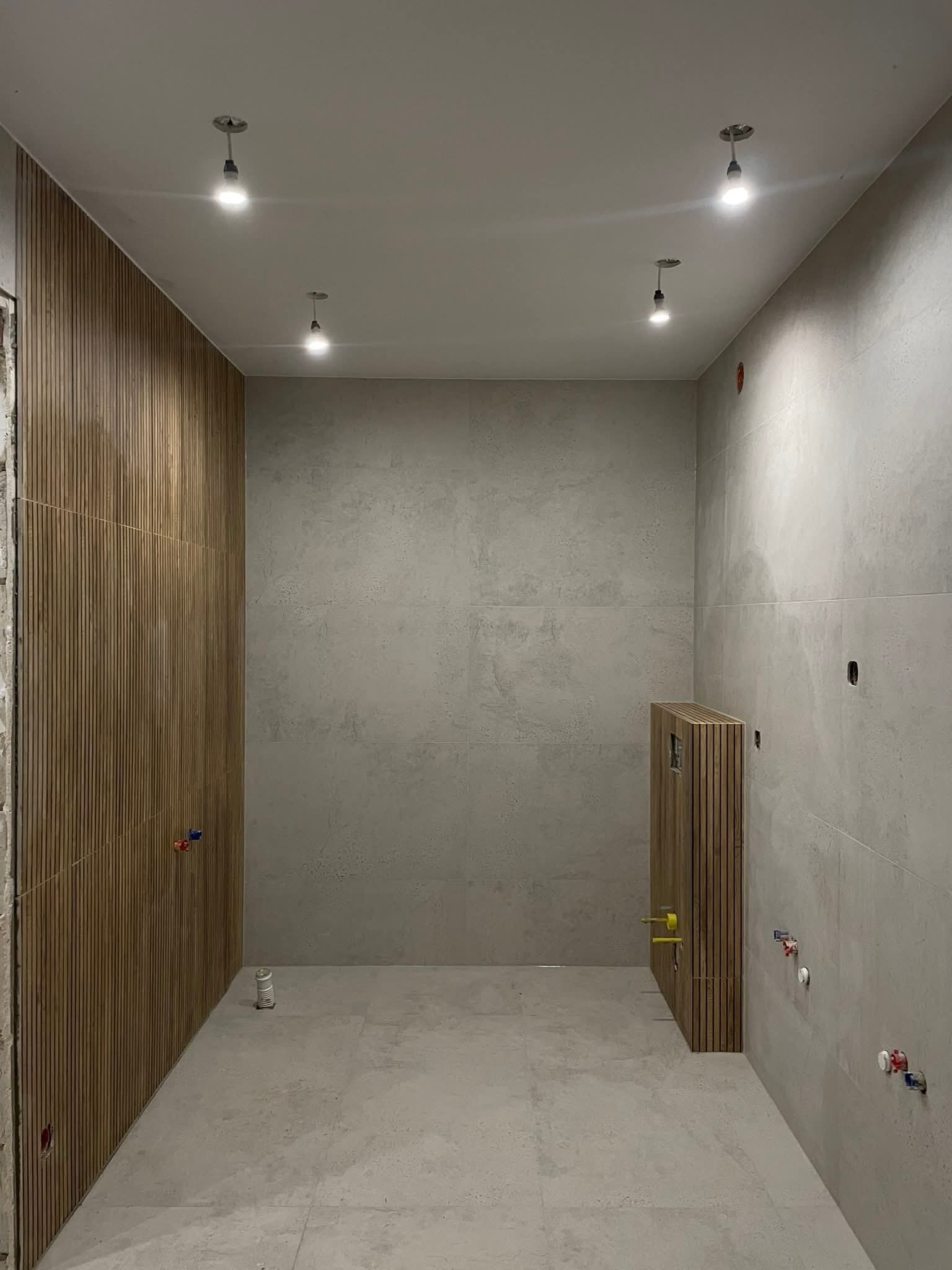 Surowa łazienka w trakcie wykańczania: drewniane panele ścienne, szare płytki, widoczne instalacje. Minimalistyczny design, oświetlenie punktowe.