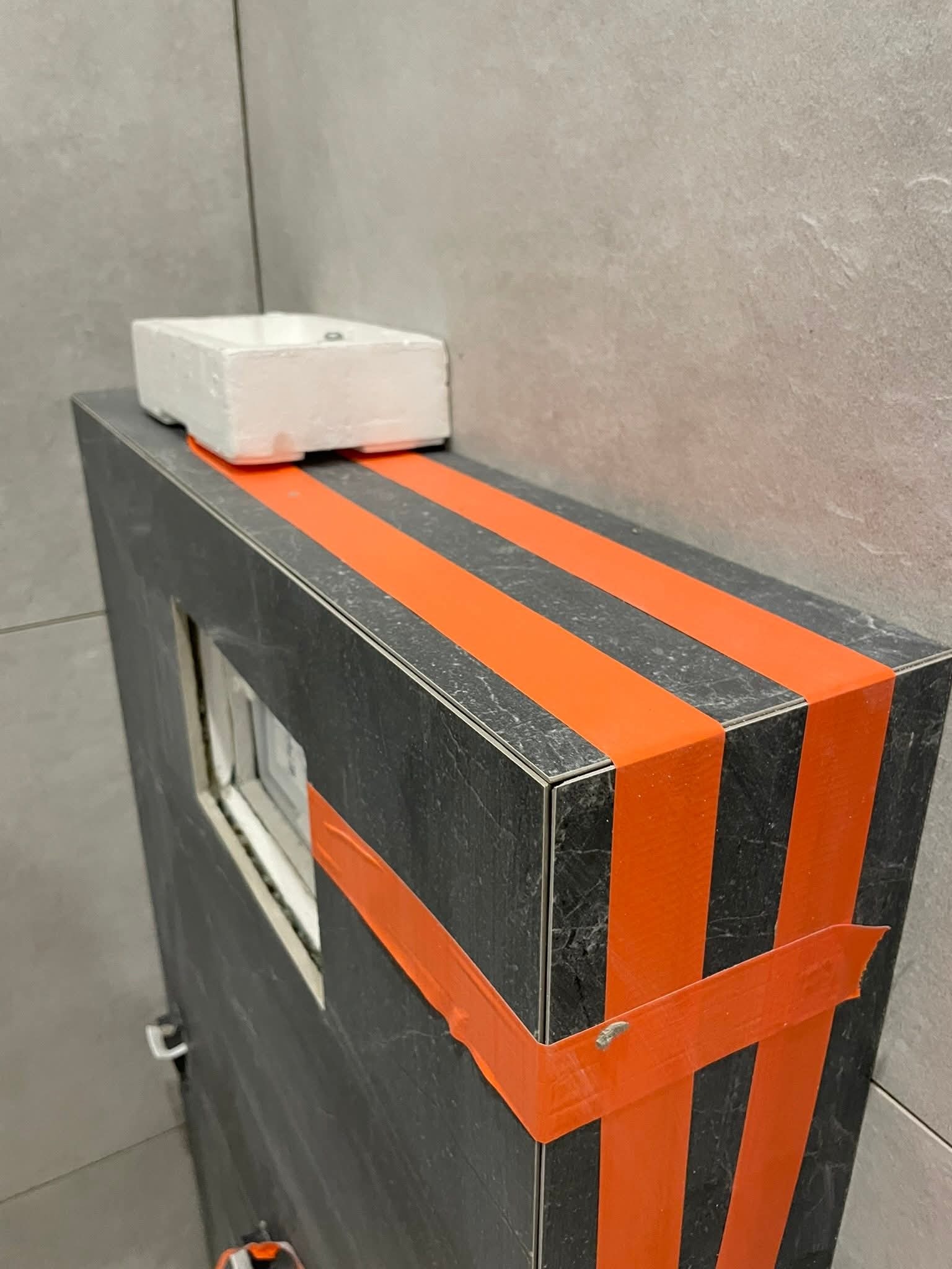 Obudowa stelaża WC z płytek imitujących kamień, zabezpieczona pomarańczową taśmą malarską. Na górze biała umywalka. Szare płytki na ścianie w tle.