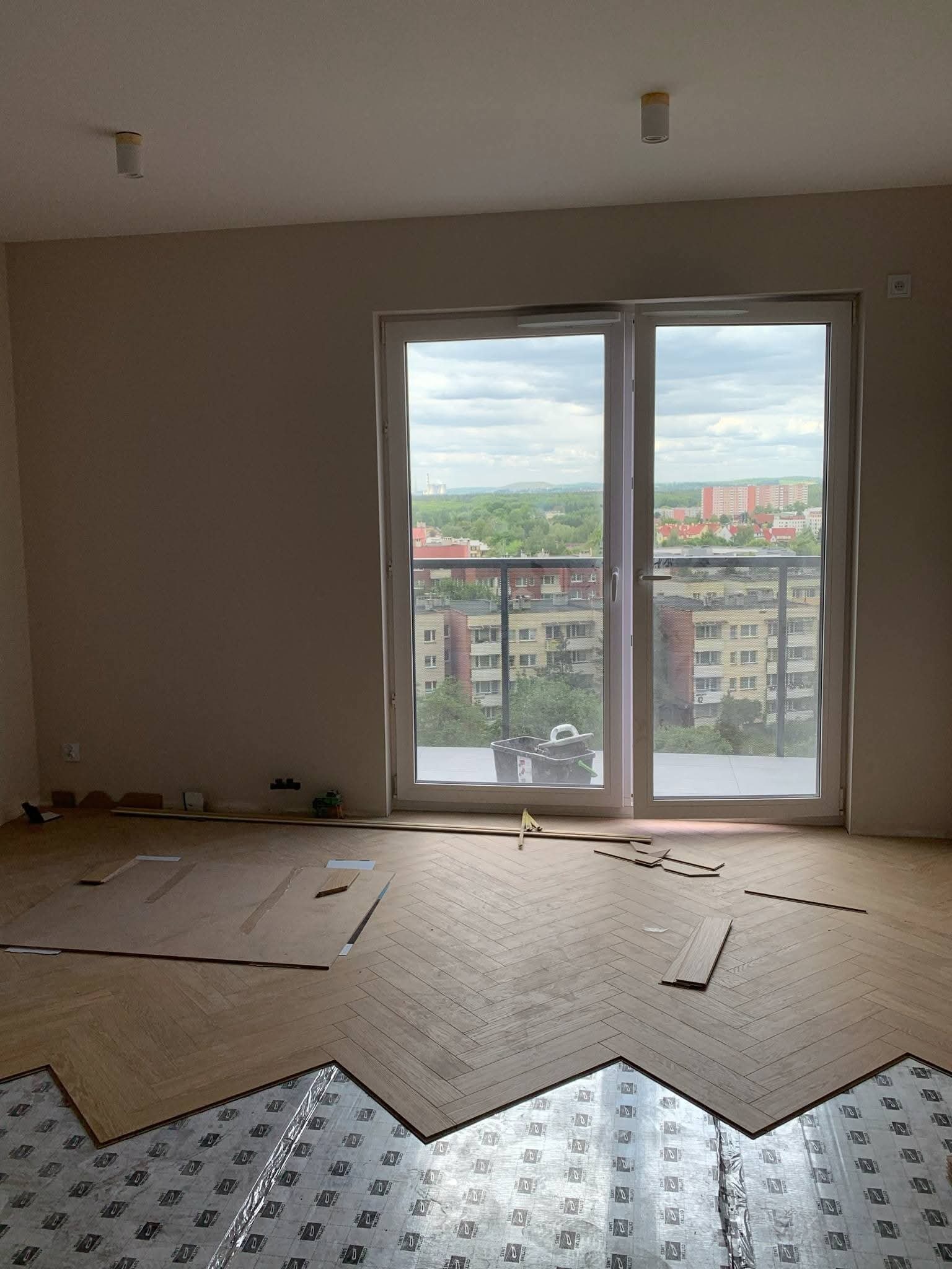 Układanie parkietu w jodełkę w jasnym pokoju z widokiem na miasto. Częściowo ułożona podłoga, widoczna folia izolacyjna. Praca w toku, naturalne światło.