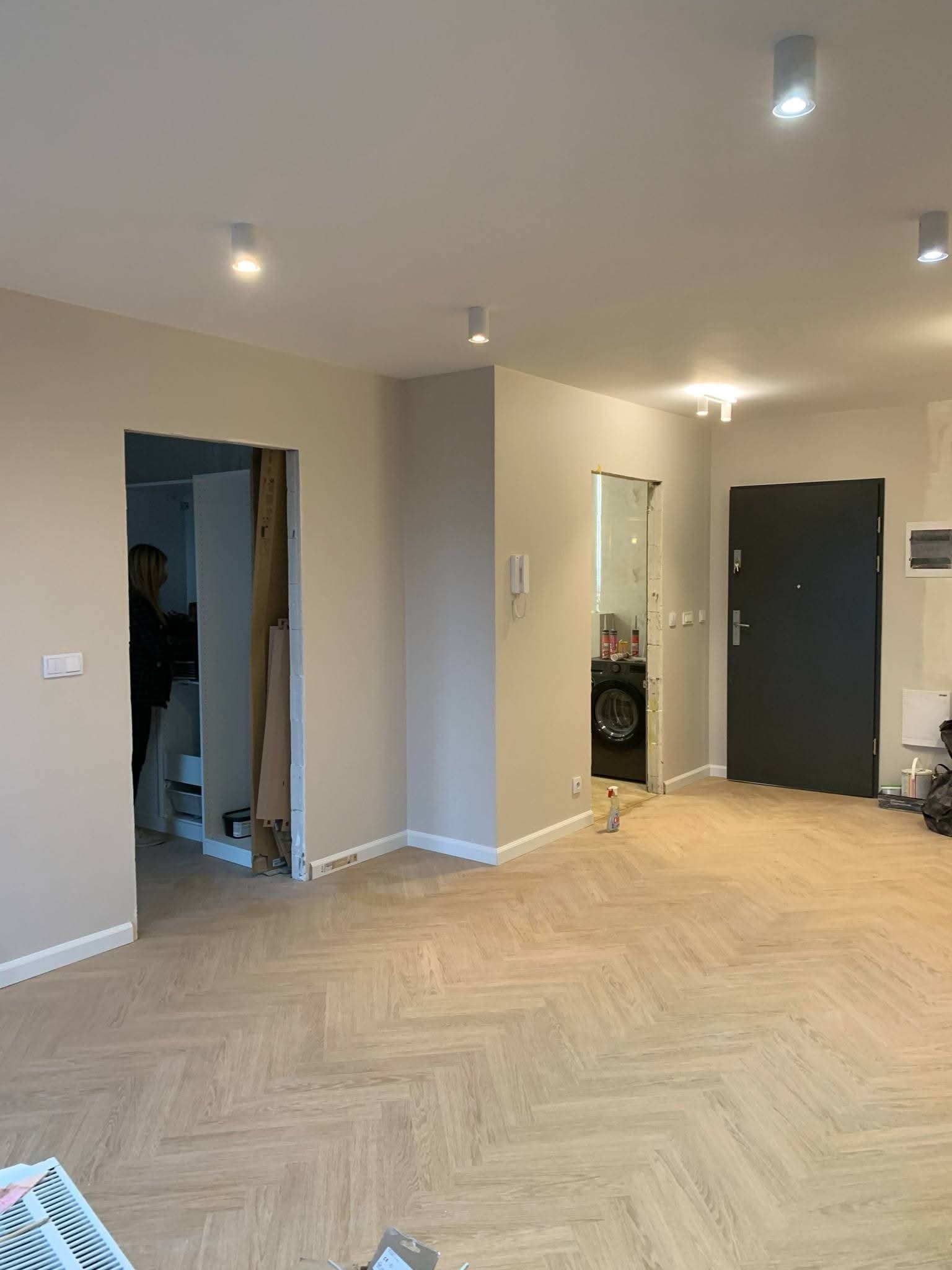 Jasne panele ułożone w jodełkę w przestronnym wnętrzu z beżowymi ścianami i widocznymi otworami drzwiowymi. W tle pralka i osoba w pomieszczeniu.