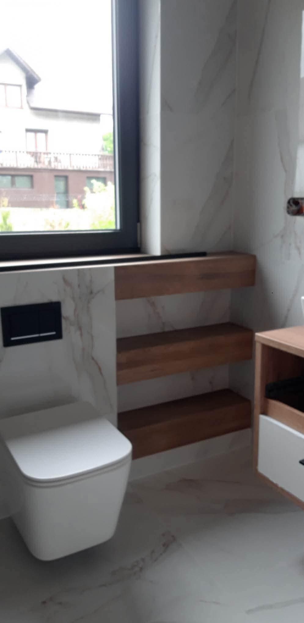 Nowoczesna łazienka z wiszącą toaletą, marmurowymi płytkami i drewnianymi półkami wnękowymi pod oknem. Minimalistyczny design z elementami naturalnego drewna.