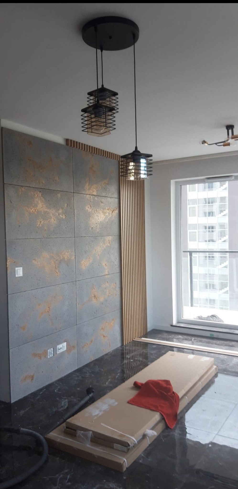 Wykończone wnętrze pokoju z dekoracyjną ścianą z paneli i płyt imitujących beton, nowoczesne lampy wiszące. Widoczne materiały budowlane na podłodze.